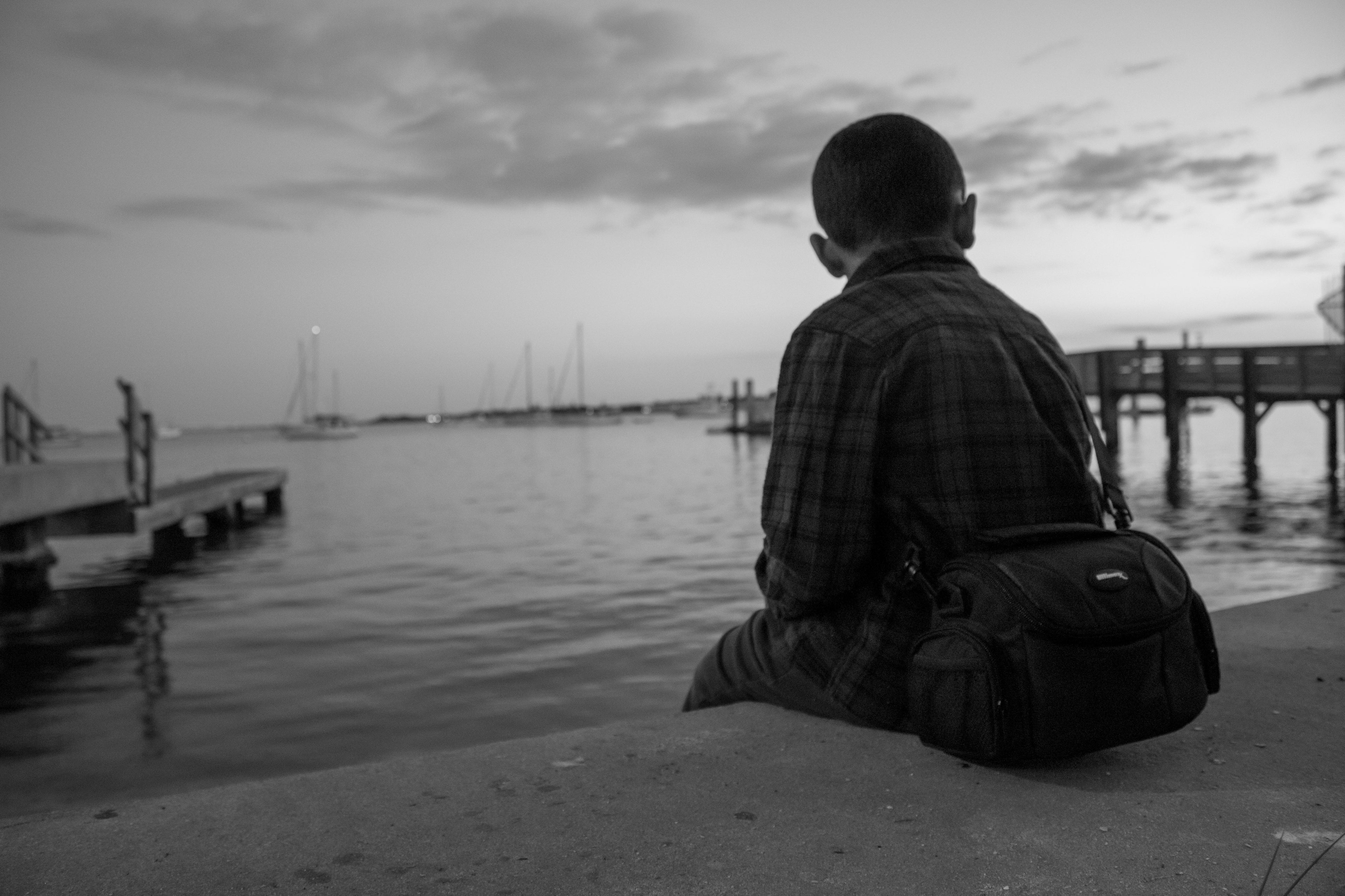 young boy sitting by a body of water with a bag, looking out at the horizon.
